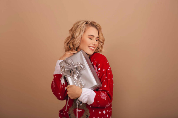 Happy smiling women in red New Year's sweater holding a silver gift box isolated on a beige background. Copy space.  - Φωτογραφία, εικόνα