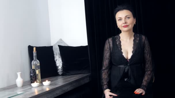Schöne junge Frau mit langen Haaren und schwarzem Peignoir und Dekolleté blickt neben Weingläsern und einer Flasche in die Kamera - Filmmaterial, Video
