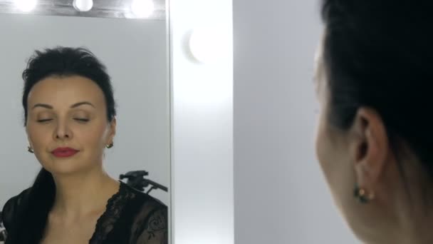 Hermosa joven vistosa con el pelo largo y en un peignoir negro y escote mira su reflejo en el espejo. - Metraje, vídeo