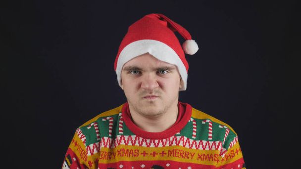 Young handsome guy shows emotion - anger, he is wearing a red New Year's sweater with New Year's drawings, and a red New Year's hat - Foto, afbeelding