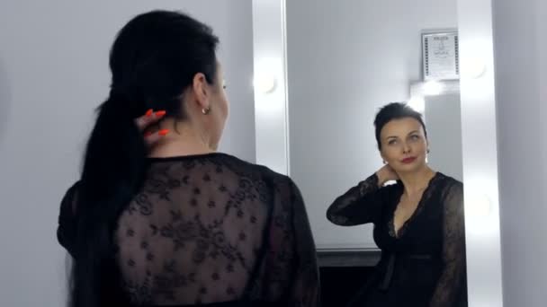 Mooie opzichtige jonge vrouw met lang haar en in een zwarte peignoir en halslijn kijkt naar haar reflectie in de spiegel. - Video