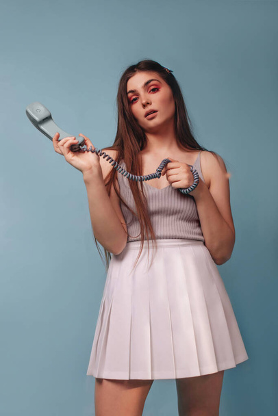 A girl with bright makeup, in a top and a white short skirt, is holding a telephone receiver isolated on a blue background. The girl is calling. - Photo, Image