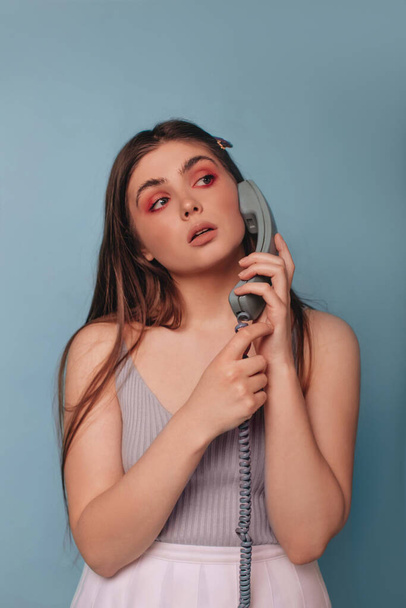 A girl with bright makeup, in a top and a white short skirt, is holding a telephone receiver isolated on a blue background. The girl is calling. - Photo, Image