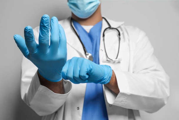 Doctor in protective mask putting on medical gloves against light grey background, closeup - Fotografie, Obrázek