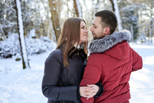 Young couple in love, enjoying winter weather outside in park forest. Brunette man in red jacket and woman in black, hugging, looking at each other. Christmas and new year holidays joy. Romantic date. - Zdjęcie, obraz