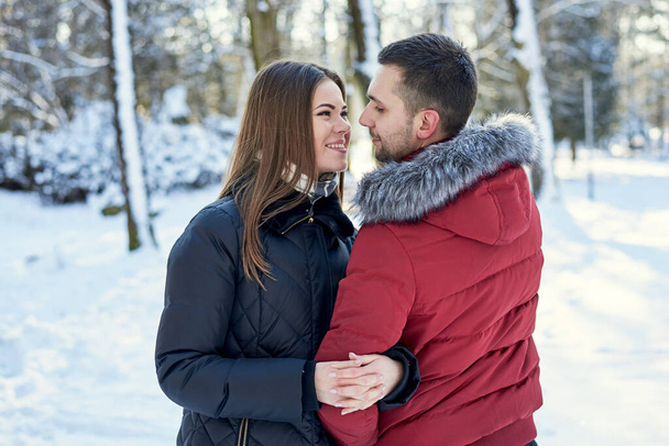 Young couple in love, enjoying winter weather outside in park forest. Brunette man in red jacket and woman in black, hugging, looking at each other. Christmas and new year holidays joy. Romantic date. - Foto, afbeelding