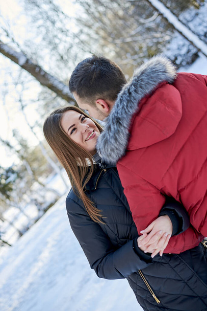 Young couple in love, enjoying winter weather outside in park forest. Brunette man in red jacket and woman in black, hugging, looking at each other. Christmas and new year holidays joy. Romantic date. - Photo, image