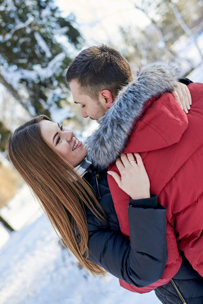 Young couple in love, enjoying winter weather outside in park forest. Brunette man in red jacket and woman in black, hugging, looking at each other. Christmas and new year holidays joy. Romantic date. - Foto, Imagem