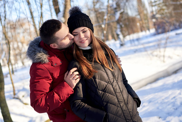 Young couple in love, enjoying winter sunny weather outside in snowy park forest. Brunette man in red jacket hugging woman in black. Christmas and new year holidays joy. Romantic date, relationship. - Fotografie, Obrázek