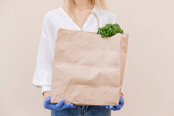 Young woman courier volunteer wearing a protective mask holds a paper bag with products, vegetables, isolated over beige background. Quarantine, coronavirus, safe food delivery. Copy space - Φωτογραφία, εικόνα