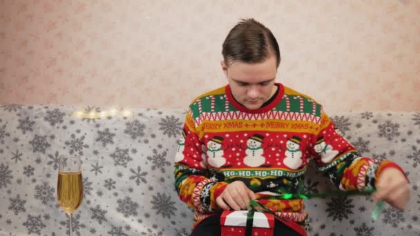 Un ragazzo con un maglione di Capodanno si siede sul divano e confeziona un regalo in una scatola rossa, confeziona giocattoli e una maschera rossa medica, accanto a un bicchiere di champagne, sceglie quale nastro avvolgere il regalo, rosso o verde. - Filmati, video