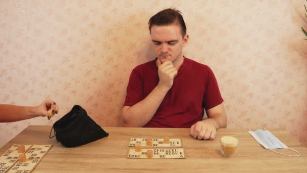 Sovyet loto oyunu, doğrulanmamış fıçılar ve numaralar, kırmızı tişörtlü bir adam loto oynar, kahve, tıbbi maske ve el dezenfektanı olan masada. - Video, Çekim