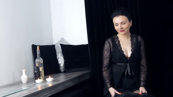Mooie jonge vrouw met lang haar en in een zwarte peignoir en halslijn kijkt naar de camera naast glazen wijn en een fles - Video