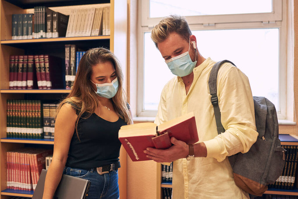 Um jovem casal usando uma máscara confira um livro da biblioteca - Foto, Imagem