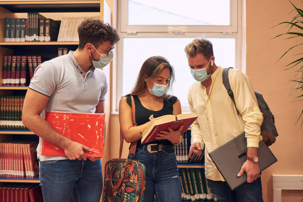Bir grup genç üniversite öğrencisi kütüphanede maske takıyor. - Fotoğraf, Görsel