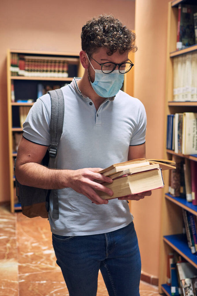 Maskeli genç bir adam üniversite kütüphanesinde kitap okuyor. - Fotoğraf, Görsel