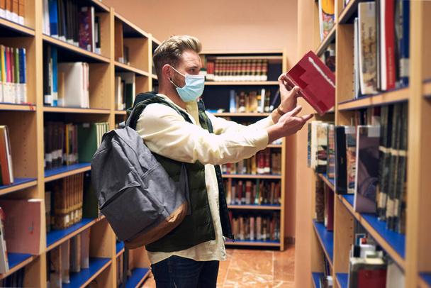 un giovane con una maschera sta prendendo un libro nella biblioteca universitaria - Foto, immagini