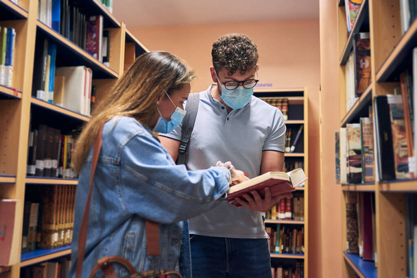 two young people wearing masks are looking at a book in the library - Foto, Imagen
