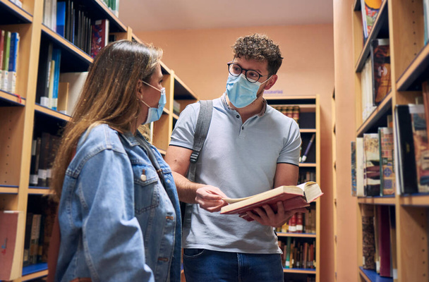 two young people wearing masks are talking in the university library - Фото, изображение