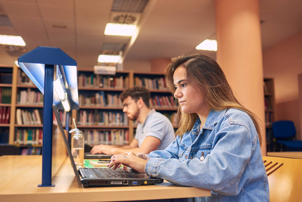 Ένα νεαρό κορίτσι χρησιμοποιεί ένα φορητό υπολογιστή ενώ σπουδάζει στη βιβλιοθήκη - Φωτογραφία, εικόνα