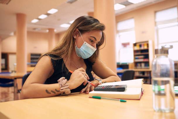 μια νεαρή γυναίκα με μια χειρουργική μάσκα που σπουδάζει στη βιβλιοθήκη - Φωτογραφία, εικόνα