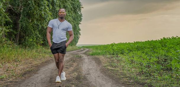 Fiatal, erős férfi egyedül sétál végig a földúton az erdő mentén, a nyári naplemente hátterében. - Fotó, kép