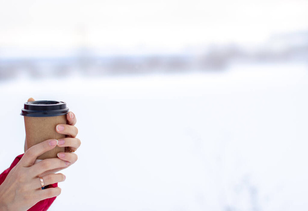 Μια νεαρή γυναίκα με κόκκινο μπουφάν το χειμώνα κρατάει ένα ποτήρι ζεστό καφέ ή τσάι. Ένας χιονισμένος χειμώνας και ένα ζεστό ρόφημα για να ζεσταθείς. - Φωτογραφία, εικόνα
