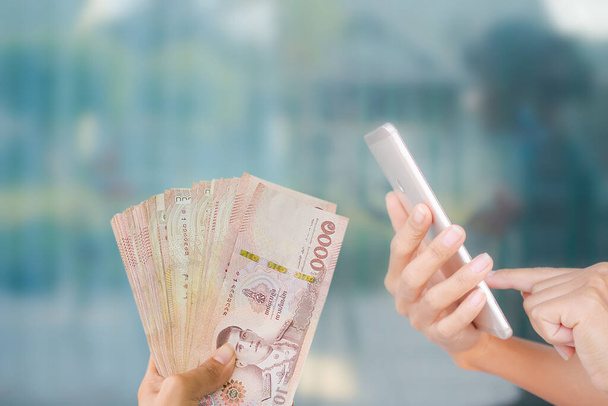 Κοντινό πλάνο με κινητό με thai χαρτονομίσματα 1000 μπατ σε μπλε φόντο. για τα συνοδευτικά άρθρα σχετικά με τις χρηματοοικονομικές συναλλαγές μέσω του τραπεζικού συστήματος του δικτύου. 2021 έτος Ξοδεύοντας χρήματα έννοια. - Φωτογραφία, εικόνα