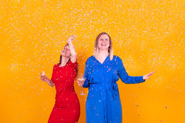 Две счастливые женщины элегантно одеты на вечеринке и празднуют вместе, держа стакан пузырьков, пока конфетти разлетаются повсюду - Фото, изображение