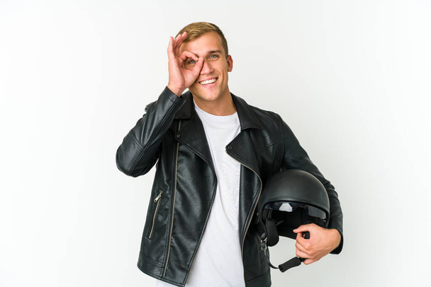 Joven hombre caucásico sosteniendo un casco de moto aislado sobre fondo blanco emocionado manteniendo el gesto bien en el ojo. - Foto, imagen