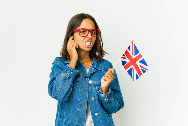 Mujer latina joven sosteniendo una bandera inglesa aislada sobre fondo blanco cubriendo las orejas con las manos. - Foto, Imagen
