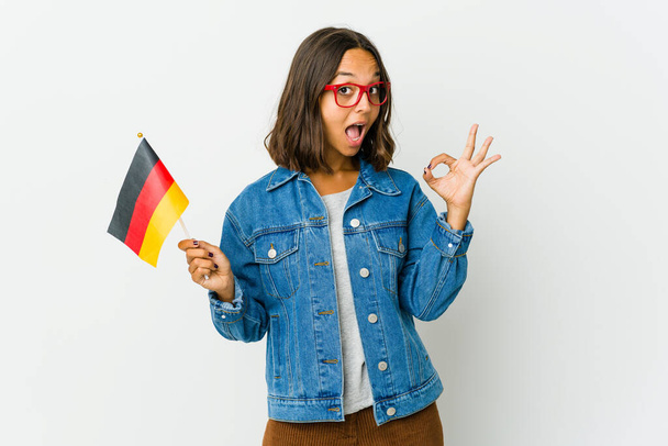 Jonge latijnse vrouw met een Duitse vlag geïsoleerd op witte achtergrond knipoogt een oog en houdt een goed gebaar met de hand. - Foto, afbeelding