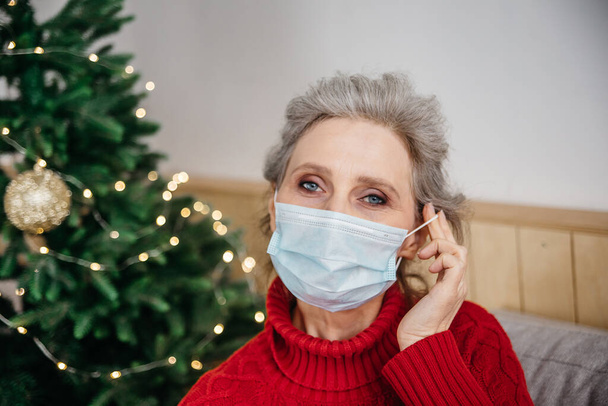 Święta Bożego Narodzenia w domach opieki podczas pandemii COVID-19. Close up portret starszej pani zakładanie maski w domu choinki na tle. - Zdjęcie, obraz