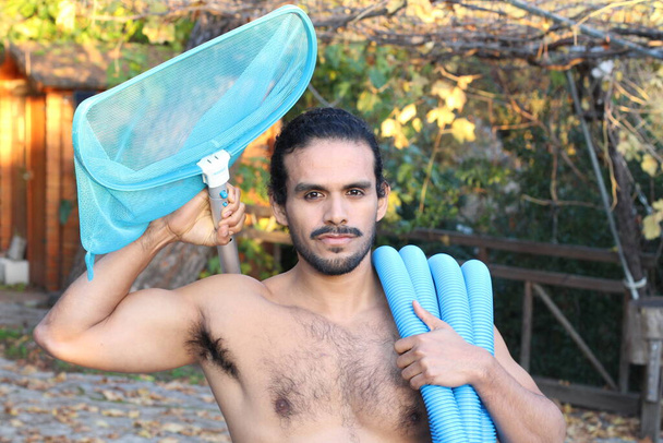 γκρο πλαν πορτρέτο του όμορφου νεαρού άνδρα με μουστάκι και μακριά μαλλιά κρατώντας δίχτυ και σωλήνα για καθαρισμό πισίνας - Φωτογραφία, εικόνα