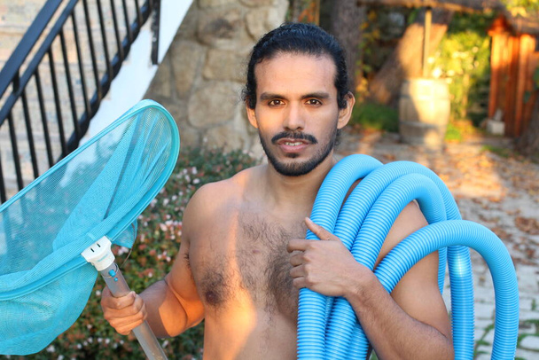 retrato de primer plano del joven guapo con bigote y pelo largo sosteniendo red y tubería para la limpieza de la piscina - Foto, Imagen