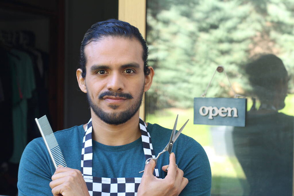 portrait of handsome Latin barber with scissors in uniform in front of door with open tab - Photo, Image