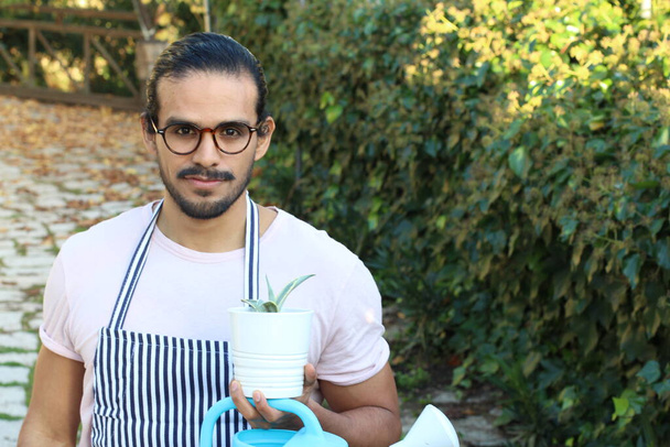 κοντινό πορτραίτο όμορφου νέου κηπουρού με ποδιά με μουστάκι και γυαλιά που κρατάει γλάστρα - Φωτογραφία, εικόνα