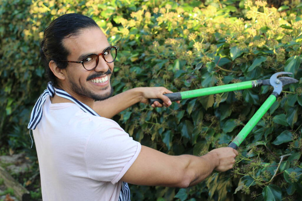 primer plano retrato de joven jardinero guapo en delantal con bigote y anteojos cortando arbustos - Foto, imagen