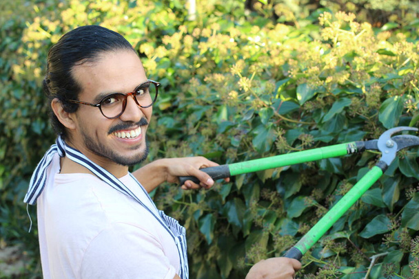 primer plano retrato de joven jardinero guapo en delantal con bigote y anteojos cortando arbustos - Foto, imagen