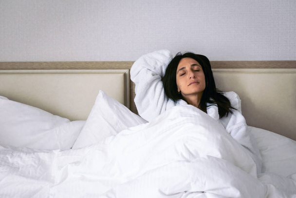 Vrouw in wit gewaad ligt vrolijk met gesloten ogen in wit bed. Relxatie op vakantie hotel en comfort concept. - Foto, afbeelding