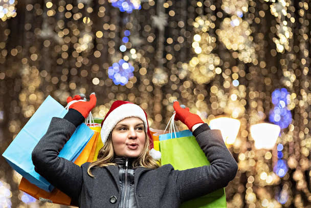 Uzun saçlı ve Noel Baba şapkalı bir kadın. Renkli kağıt torbalarla alışveriş yapan bir mağazanın penceresinin yanında. Yeni yıl alışverişi. - Fotoğraf, Görsel