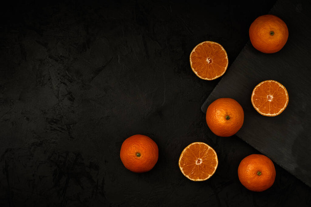 ripe whole and sliced tangerines lie on a dark concrete background and a stone cutting board. top view. artistic fruity citrus concept with copy space - Φωτογραφία, εικόνα