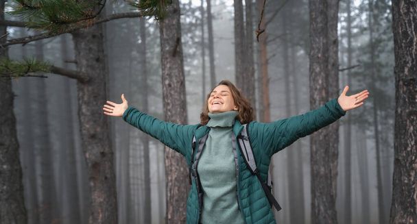 Young happy woman traveler in a green jacket with backpack in a foggy pine forest enjoying a walk hugging the world - Foto, afbeelding