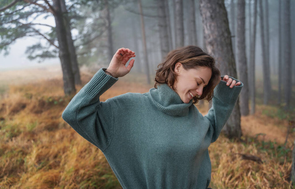 Happy woman in warm cozy green sweater enjoying nature in beautiful foggy pine forest - Photo, image