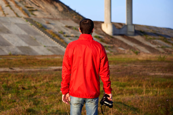 Ένας άντρας με κόκκινο μπουφάν και κάμερα στέκεται κοντά στη γέφυρα νωρίς το πρωί του φθινοπώρου, πίσω όψη. - Φωτογραφία, εικόνα