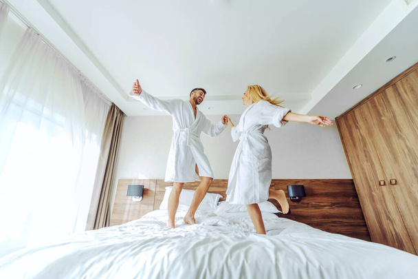 Ein fröhliches Paar mittleren Alters in Bademänteln feierte am Wochenende in einem Hotel seinen Jahrestag. Sie springen auf das Bett. - Foto, Bild