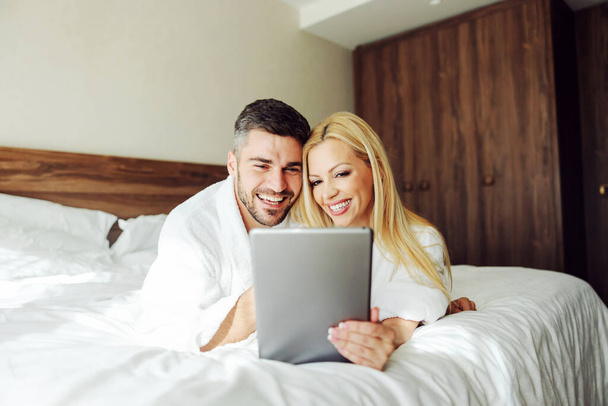 Een verliefd stel van middelbare leeftijd dat een tablet gebruikt terwijl ze op een huwelijksreis op bed liggen. Hotel appartement interieur. - Foto, afbeelding