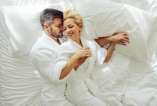 Κάτοψη ενός μεσήλικου ζευγαριού ερωτευμένου με μπουρνούζια στο μήνα του μέλιτος ξαπλωμένο στο κρεβάτι και αγκαλιασμένο. - Φωτογραφία, εικόνα