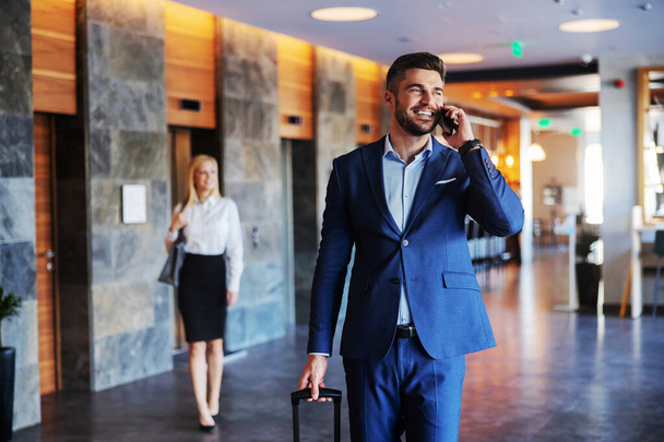 Un uomo sorridente di mezza età vestito con abiti formali che cammina nell'atrio di un hotel di lusso e parla al telefono. Sta tirando fuori la valigia.. - Foto, immagini
