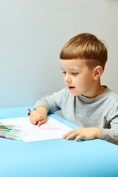 Sarı saçlı sevimli küçük çocuk evde renkli kalemler çiziyor. Mavi masada çizer. - Fotoğraf, Görsel